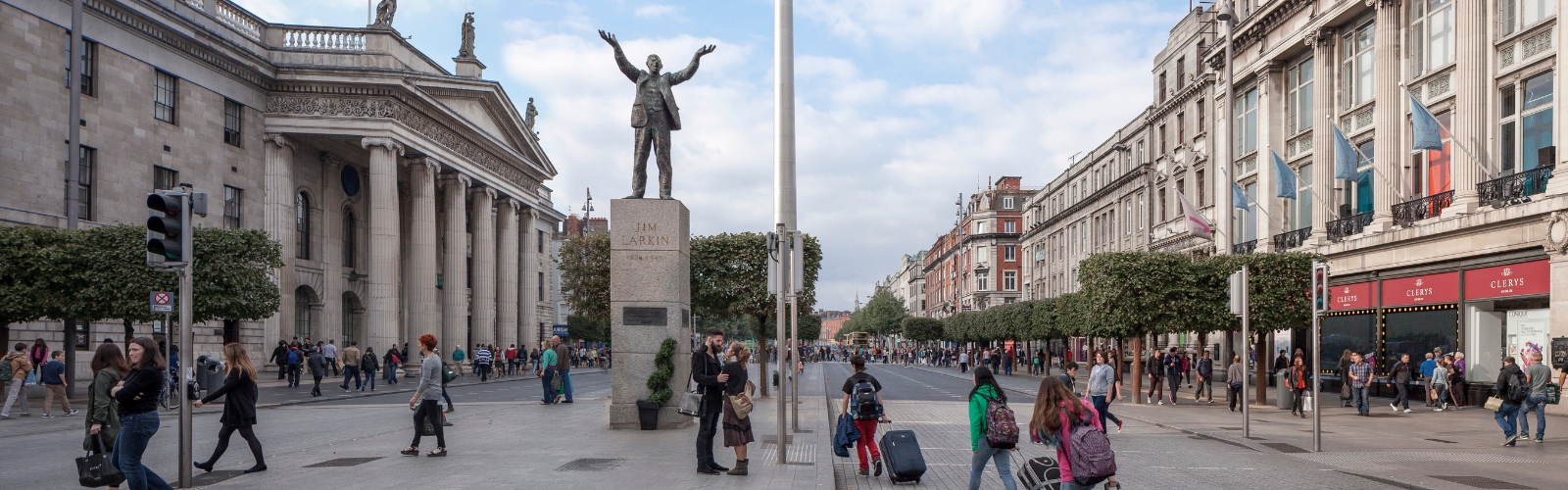 A bronze statue of a man with his arms in the air is situated in the middle of a busy street next to a stone building with many pillars. 